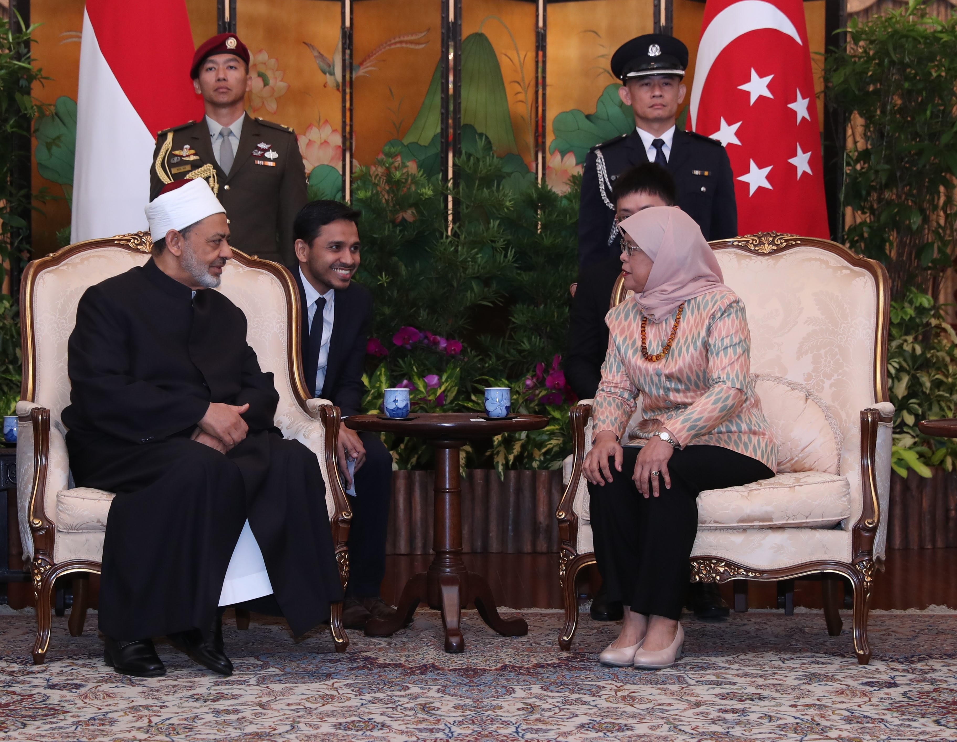 Two people seated in ornate chairs, talking; flags of Indonesia and Singapore in background.